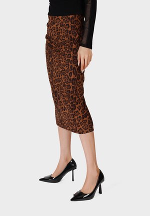 Donna con gonna marrone stampata leopardata fino al ginocchio, tacchi alti neri e top nero a maniche lunghe, in piedi davanti a uno sfondo semplice.