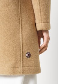 Beiger Wollmantel mit strukturierter Stoffqualität, geradem Schnitt und einem runden, dunkelroten Logo-Knopf an der unteren Seite.