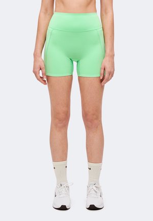 Femme portant un short moulant taille haute vert vif, des chaussettes blanches et des chaussures de sport blanches, debout devant un fond uni.