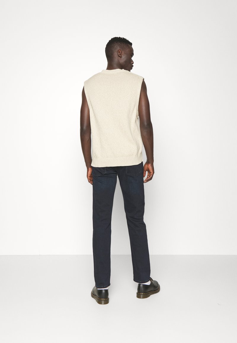 Beige gestrickter ärmelloser Pullover mit Rippenbesatz, kombiniert mit dunkelblauen Slim-Fit-Jeans und schwarzen Schuhen. Glatte Textur, schlichtes Design.