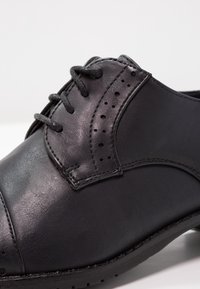 Chaussure habillée en cuir noir avec des détails brogués sur la rangée d'œillets et le bout, dotée de lacets noirs et de coutures surpiquées.