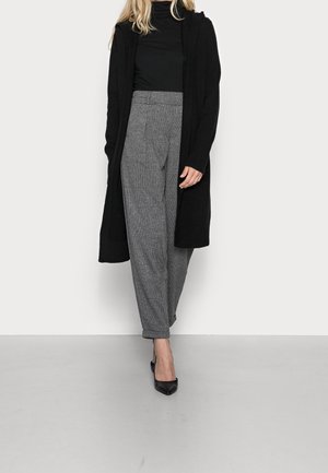 Kobieta w czarnym długim kardiganie na czarnym golfie, szare spodnie z wysokim stanem i szerokimi nogawkami oraz czarne szpilki z ostrym noskiem stoi na szarym podłożu.