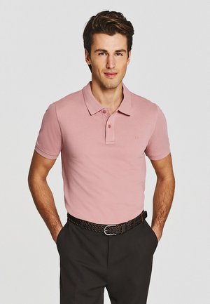 Mężczyzna noszący różową koszulkę polo z krótkim rękawem, wpuszczoną do czarnych spodni z plecionym paskiem, stoi z rękami w kieszeniach.