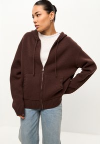 Sudadera marrón con cremallera, con dobladillo y puños acanalados, que presenta una capucha y dos bolsillos frontales, combinada con jeans azul claro.