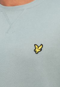 Felpa leggera azzurra a girocollo realizzata in tessuto morbido, con un piccolo logo di un'aquila nera e gialla sul petto. Dettagli di cucitura visibili.