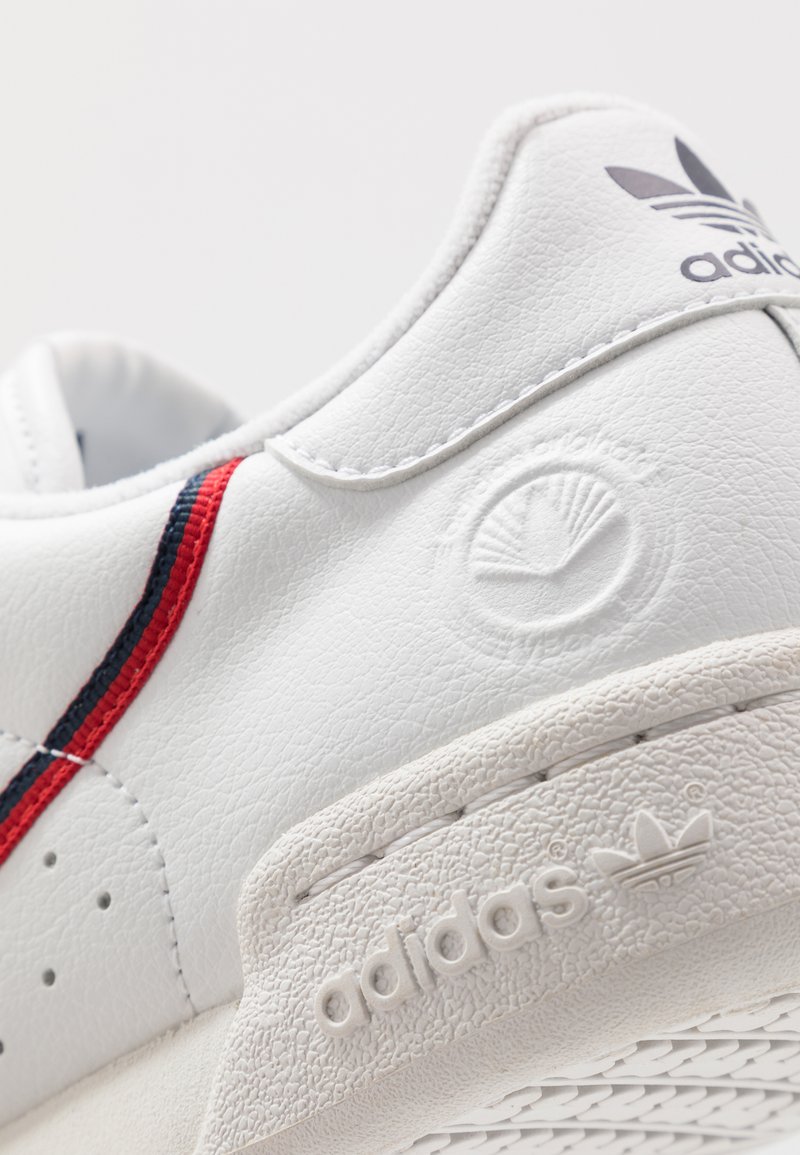 White leather sneaker featuring red and navy stripes, embossed logo, textured sole, and distinct stitching details.