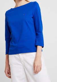 Blå bomullströja med lång ärm och båthals. Har en liten logotyp på bröstet, kombinerat med vita byxor med vida ben. S Regelbunden textur.