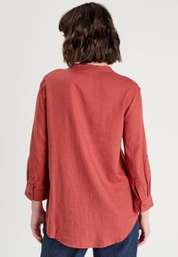 Blouse en lin rouge avec des manches trois-quarts, des poignets boutonnés, un ourlet arrondi et une coupe décontractée, mettant en valeur une surface texturée.