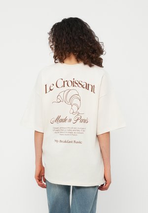 Femme aux cheveux bouclés portant un T-shirt blanc surdimensionné avec un graphique de croissant marron et un texte en français au dos.