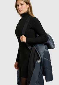 Fekete bordázott kötött ruha magas nyakkal; a modell egy sötétkék, fényes anyagú, logós pánttal ellátott puffert tart.