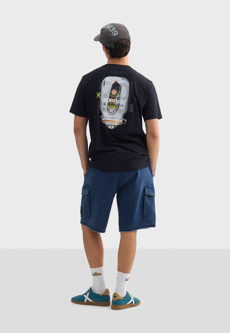 Černé tričko s potiskem na zadní straně, spárované s modrými cargo šortkami, bílými ponožkami a tyrkysovými teniskami s hnědými akcenty.