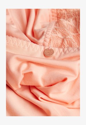 Pfirsichfarbener Stoff mit weicher Textur, der an der oberen Kante aufwändige Spitzenverzierungen und ein kleines Metallschild mit der Aufschrift "cache coeurs" aufweist.