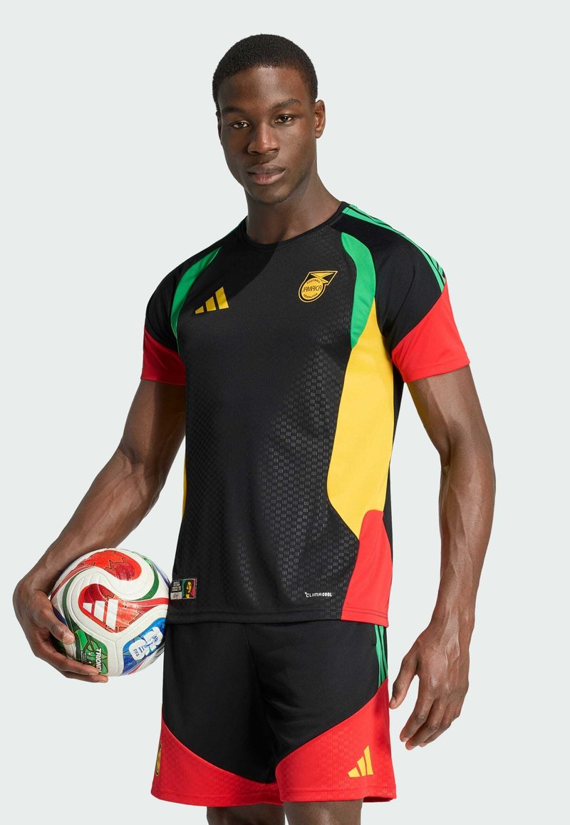 Uomo in divisa da calcio nera, rossa, gialla e verde che tiene un pallone da calcio Adidas multicolore, in piedi davanti a uno sfondo semplice.