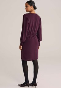 Donna con i capelli scuri raccolti in un bun, indossa un vestito a maniche lunghe color prugna, collant neri e tacchi a punta scuri, in piedi davanti a uno sfondo beige.