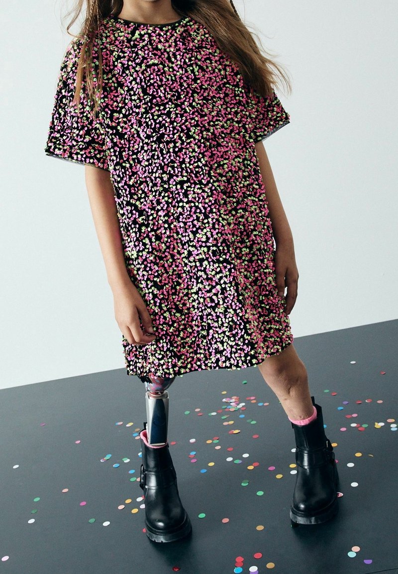Jeune fille portant une robe noire avec des motifs floraux roses et verts, une jambe prothétique et des bottes noires, debout sur un sol couvert de confettis.