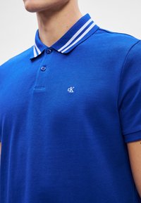 Verebek színű, textúrált anyagból készült póló, három gombos gallérral, fehér csíkozott nyakkal és kicsi logóval a bal mellen.