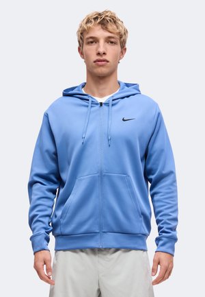 Jeune homme aux cheveux blonds courts et bouclés, portant un sweat à capuche zippé bleu clair avec un logo Nike sur la poitrine.