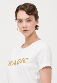 Hvit T-skjorte med korte ermer, dekorert med glitterende gullbokstaver som sier "MAGIC" på forsiden. Mykt stoff med rund hals og rullede ermer.