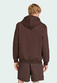 Hnedá mikina s veľkou, nastaviteľnou kapucňou, rebrovanými manžetami a lemom, zobrazená zozadu. Nosená s rovnako hnedými šortkami.