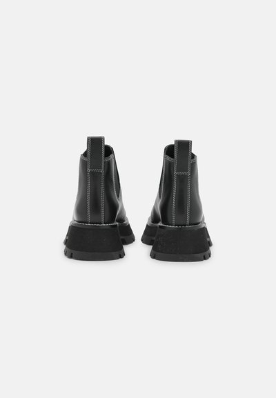 Μαύρες δερμάτινες μποτίνες μέχρι τον αστράγαλο με χοντρή σόλα, διαθέτοντας αντίθεση άσπρη ραφή και λαβές στο πίσω μέρος για εύκολη φορεσιά.