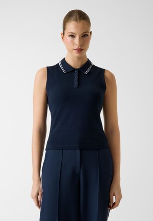 Donna che indossa una polo senza maniche blu navy con colletto a righe bianche e pantaloni plissettati blu navy abbinati, in piedi davanti a uno sfondo semplice.