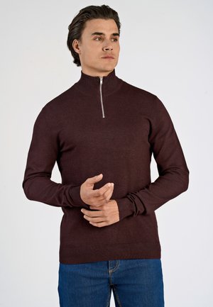 Pulover tricotat bordeaux cu guler înalt și închidere cu fermoar, design ajustat, manșete și tiv cu milă, purtat cu blugi albaștri.