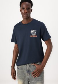 Camiseta de algodón azul marino con un gráfico de un perro con una gorra y el texto "LOS QUE HACEN" en naranja. Diseño sencillo de cuello redondo.