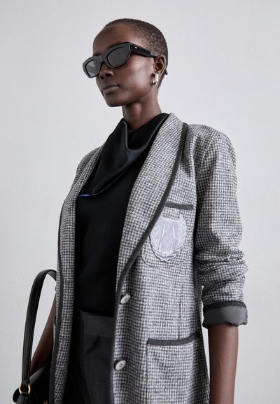 Blazer en pied-de-poule gris avec un blason, haut noir et pantalon gris. Des lunettes de soleil noires et des boucles d'oreilles texturées complètent le look.