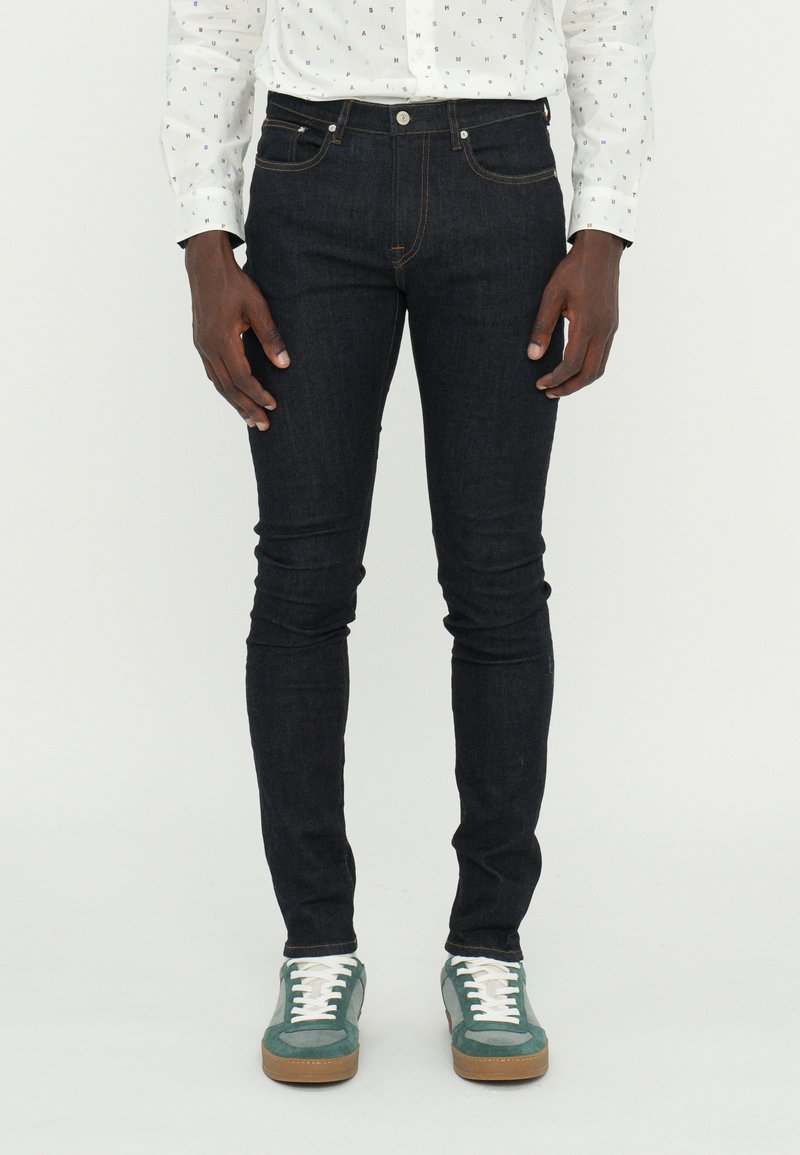 Jean skinny en denim bleu foncé avec un design à cinq poches, coutures contrastées, portés avec des baskets vertes et grises et une chemise à motifs.