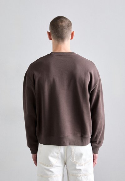 Sweatshirt en coton brun avec un col côtelé, coupe large et coutures d'épaule tombantes, associé à un pantalon blanc. Pas de motifs ou d'accents notables.