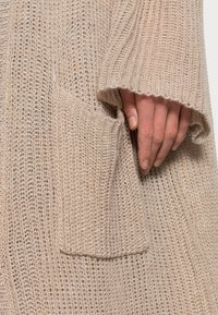 Maglione beige lavorato a maglia con texture a trama aperta, grande tasca e maniche ampie per una vestibilità rilassata. Filato morbido con un motivo di punto largo.