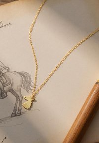 Goldene Halskette mit einer dünnen Kette und einem Pferdekopf-Anhänger. Der Anhänger weist minimale Details und eine glatte Oberfläche auf. Der Hintergrund enthält Papier und einen Bleistift.