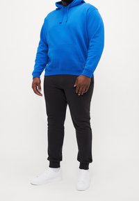 Uomo che indossa una felpa blu brillante, pantaloni jogger neri e sneakers bianche, in piedi contro uno sfondo bianco semplice.