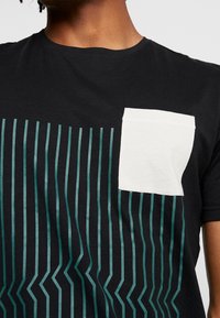 Sort T-shirt med teal lodrette striber og zigzag mønster, der har en hvid lomme på venstre bryst og en klassisk rund halsudskæring.