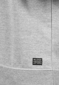 Felpa grigia con una texture liscia, orlo a coste e un'etichetta nera che riporta "BLEND" e "GLOBAL JEANMAKER" sul lato inferiore.
