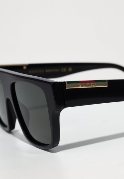 Čierne slnečné okuliare s hrubým rámom, tmavými šošovkami a zlatými detailmi. Obsahujú zelené a červené akcenty na stranách s menovkou "GUCCI".