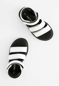 Svarta sandaler med tre vita kardborreband, med en mjuk inredning och platt sula. Varumärket "NEXT" syns på inläggen.