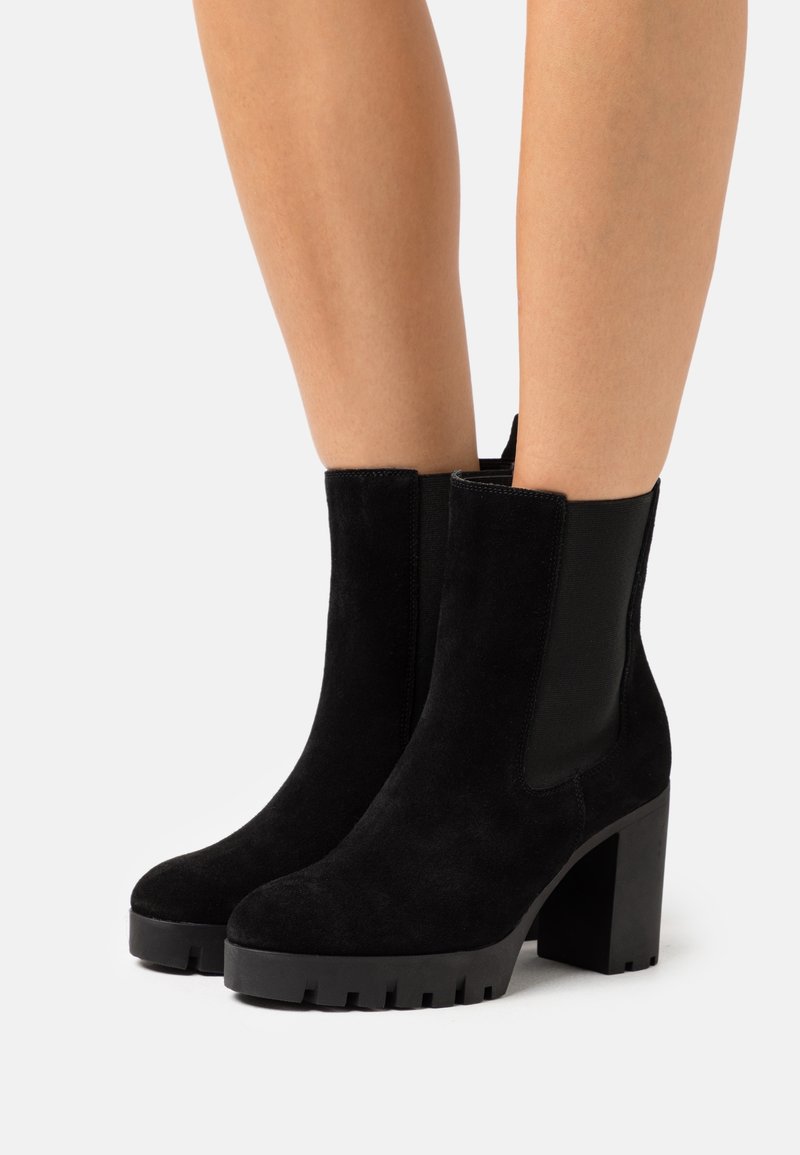 Bottines en daim noir avec talons hauts épais et panneaux latéraux élastiques portées sur des jambes nues contre un fond blanc.