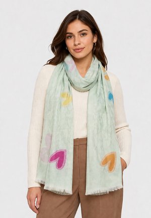 Woman wearing a light green scarf with multicolored heart patterns over a cream sweater and brown pants, standing against a plain background.