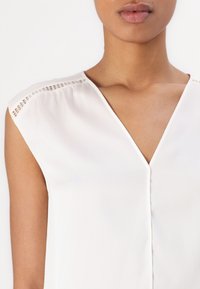 Blouse blanche sans manches avec un col en V, ornée de clous décoratifs sur la couture d'épaule et une texture de tissu lisse et léger.