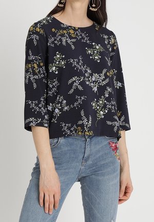 Kvinde iført en marineblå top med blomsterprint og 3/4-lange ærmer samt lyseblå jeans med rødt blomsterbroderi på lommen.