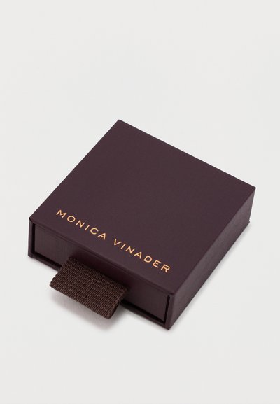 Tumši brūns kvadrātveida rotaslietu kutija ar pacelšanas cilpu un „Monica Vinader” zelta tekstu uz vāka, novietota uz baltas virsmas.