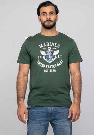 Homme aux cheveux noirs et à la barbe portant un t-shirt vert foncé "Marines United States Navy" et un jean bleu, debout devant un fond uni.