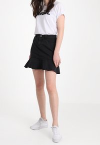 Svart denimkjol med utsvängd fåll, bärs med en vit grafisk T-shirt och vita sneakers. Har en knappstängning och fickor.