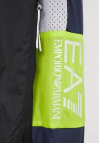 Black and navy windbreaker with green accent. Features a zipper, mesh panel, and "EMPORIO ARMANI" logo in white. Textured fabric.
