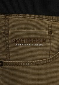 Oznaka hlača PME Legend s smešnim šavovima na teksturiranom maslinasto zelenom materijalu, s jasnim ispisom "PME LEGEND" i "AMERIKANSKA KLASIČNA".