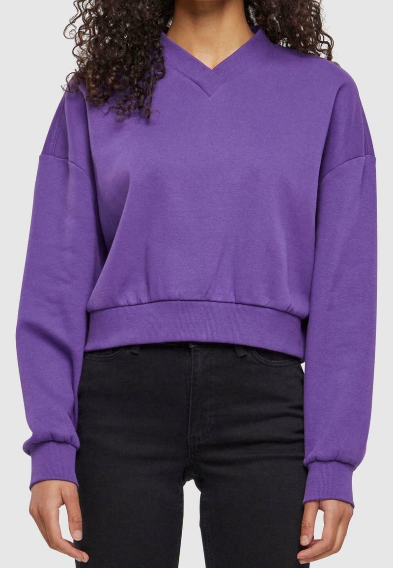 Personne portant un sweat court violet à col en V avec manches longues et un jean noir ajusté, debout devant un fond uni.