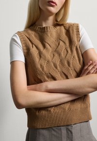 Brown knit sleeveless sweater with a textured cable design and ribbed neckline and hem, worn over a white fitted top.