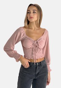 Top corto rosa con corpo arricciato e chiusura a fiocco, caratterizzato da maniche lunghe e puffate con polsini elastici. Abbinato a jeans a vita alta in denim scuro.