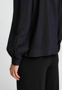 Blusa nera con tessuto testurizzato, vestibilità ampia, maniche voluminose e polsini abbottonati. Abbinata a pantaloni neri su misura, che mostrano una trama liscia.